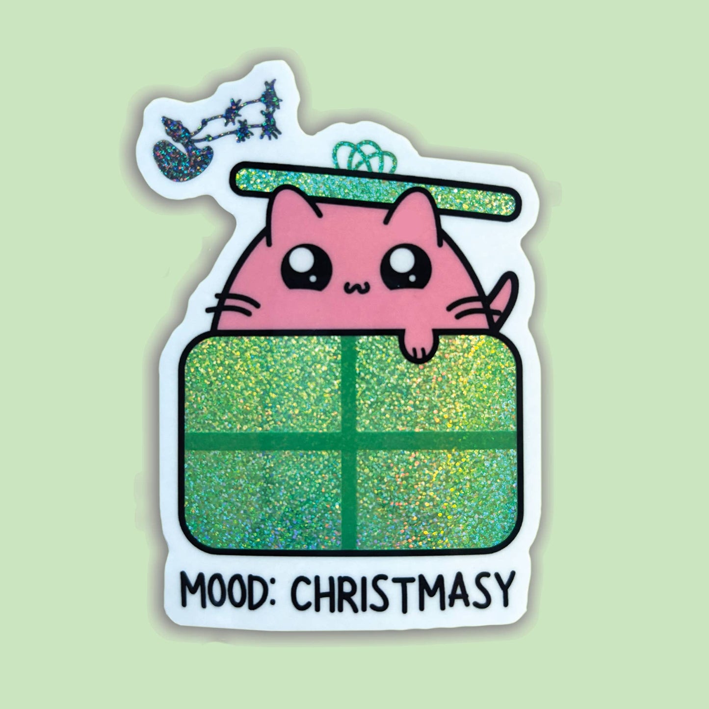 A die-cut sticker featuring a cute pink cartoon cat peeking over a glittery green Christmas gift box, with a small sleigh and stars above and the text 'MOOD: CHRISTSMASY' at the bottom, shown on a pale green background.
