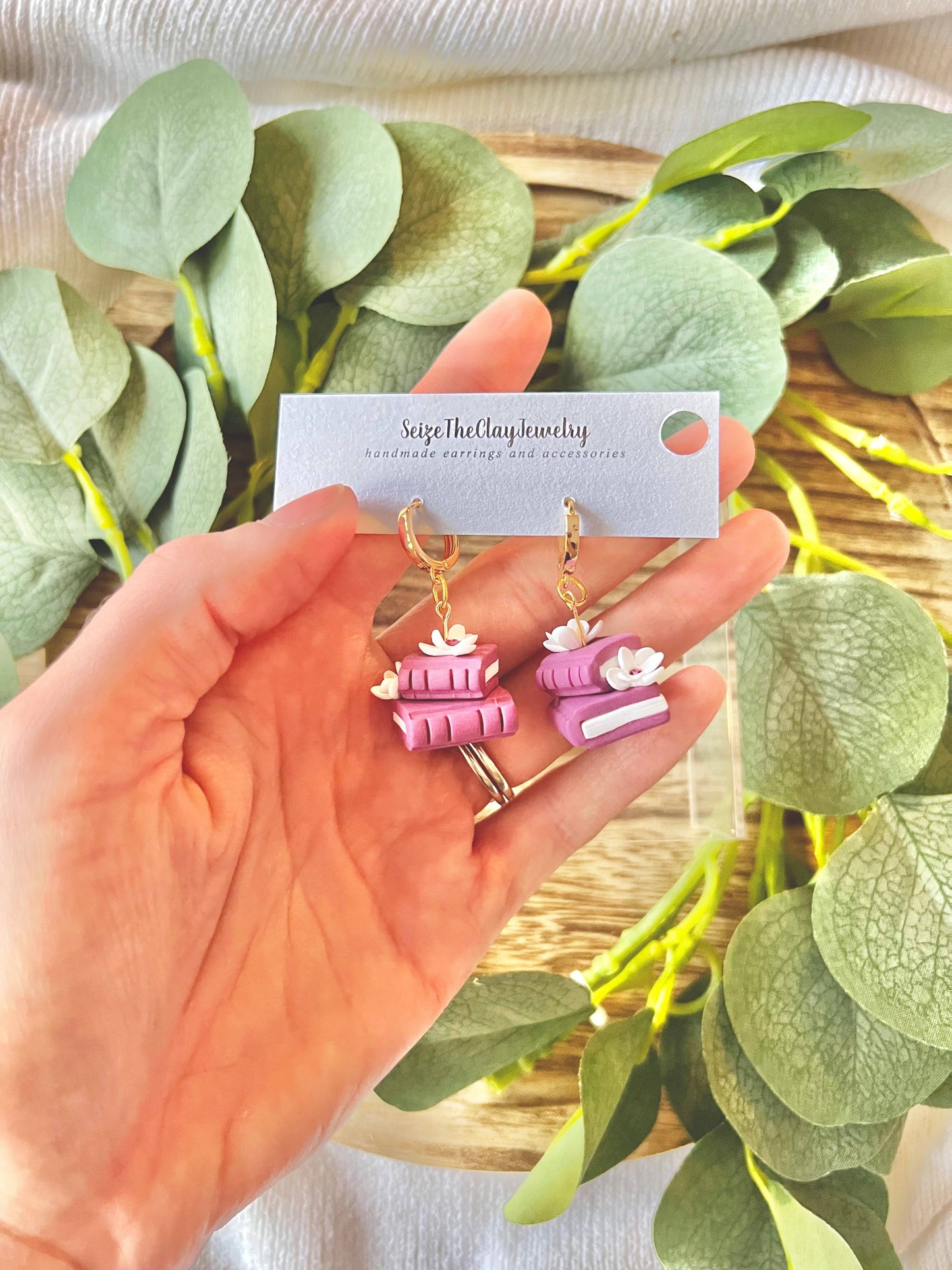 Hand holding a card with earrings featuring stacked books against a leafy background