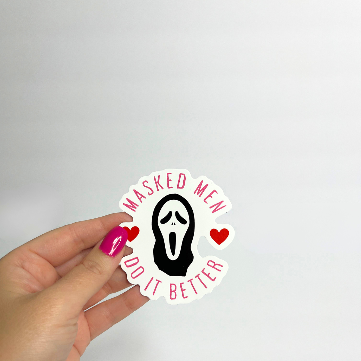 Sticker with 'Masked Men Do It Better' text held by a hand on a light gray background