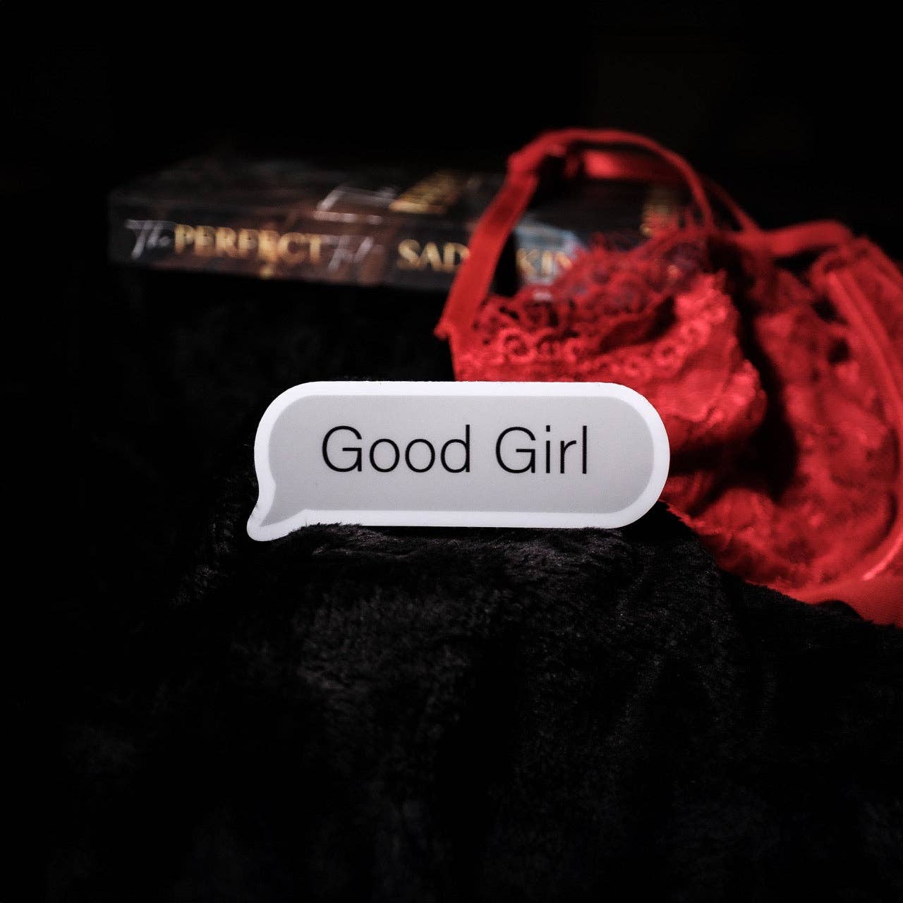 Text message bubble with 'Good Girl' on a dark background with red object and book.