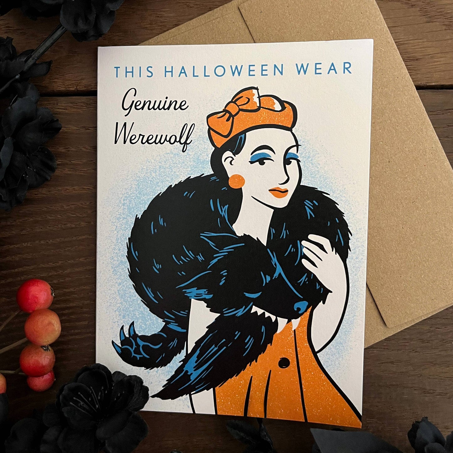 A Halloween-themed greeting card featuring a vintage-style illustration of a woman in a black werewolf fur coat and orange dress, with the text 'THIS HALLOWEEN WEAR Genuine Werewolf'. The card is displayed on a brown kraft envelope over a dark wood surface with black flowers and small red berries as props.