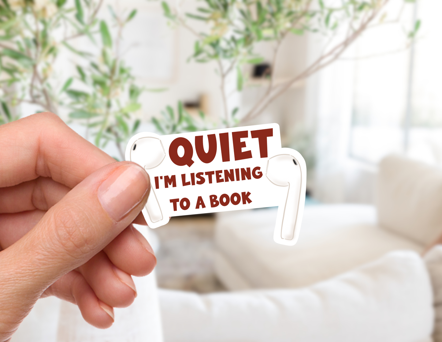 A hand holds a die-cut sticker shaped like a book page with the text 'QUIET I'M LISTENING TO A BOOK' in dark red lettering, set against a softly blurred living room background with a sofa and plants.