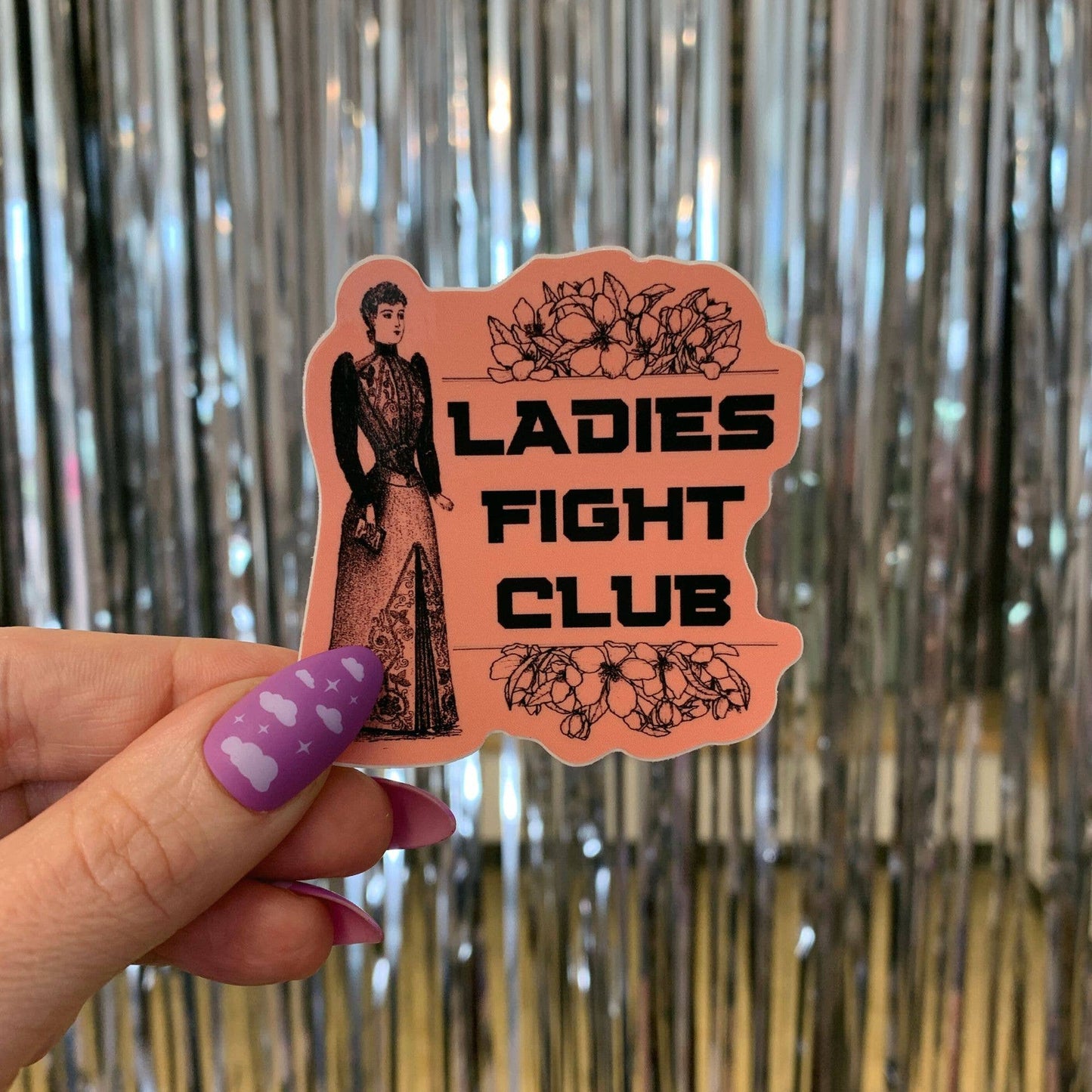 Sticker with 'Ladies Fight Club' design held by a hand against a metallic curtain background