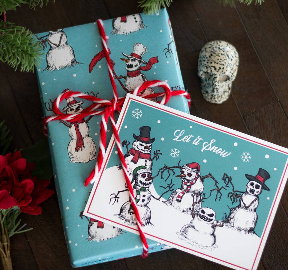 A wrapped gift box and matching greeting card featuring a winter snowman theme, tied with a red-and-white candy cane ribbon on a dark wooden surface with decorative greenery and a skull prop.