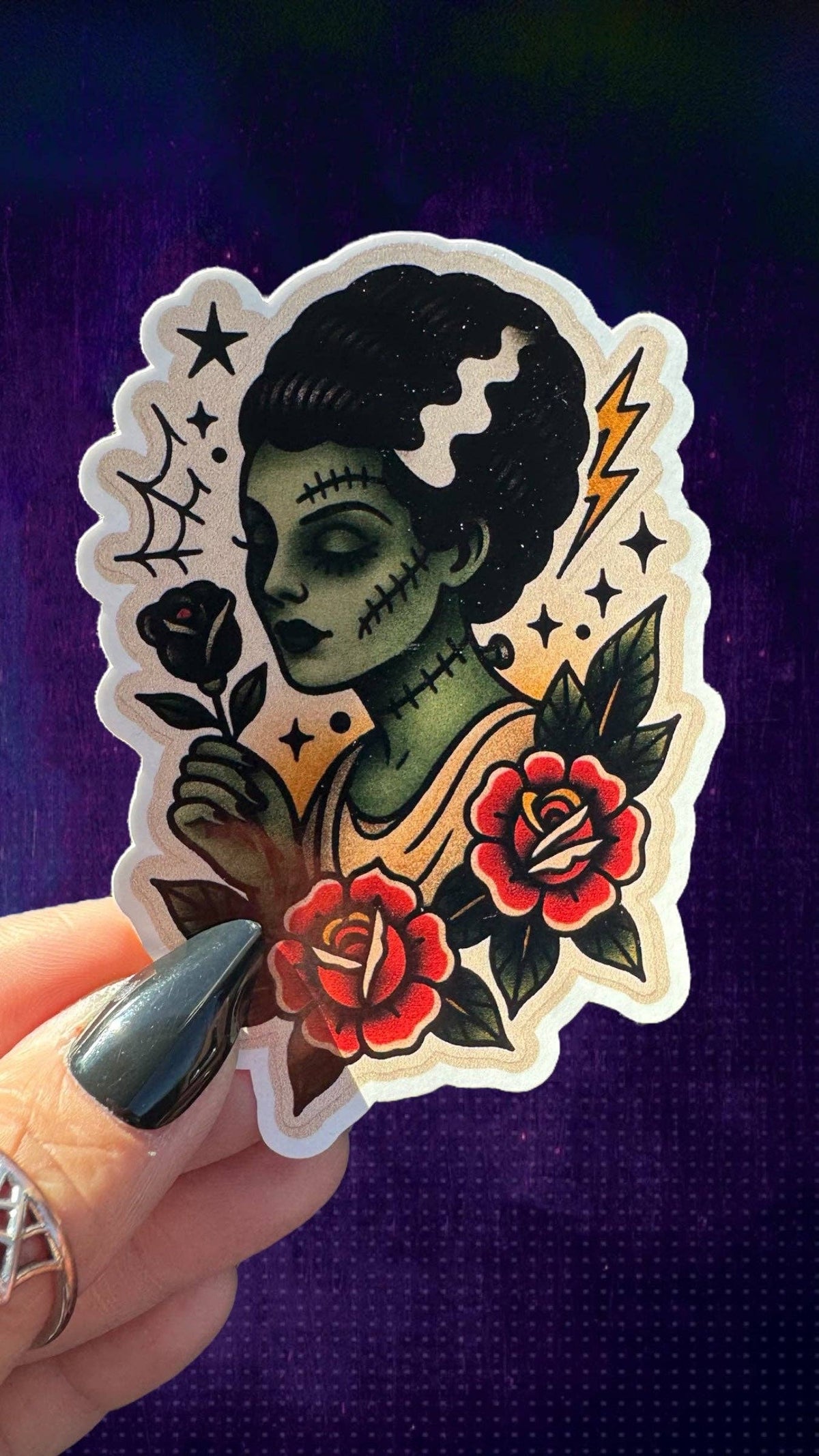 A hand with dark green nail polish holds a die-cut sticker featuring a traditional tattoo-style illustration of a green-skinned woman with black hair, red roses, and a lightning bolt, set against a dark purple textured background.