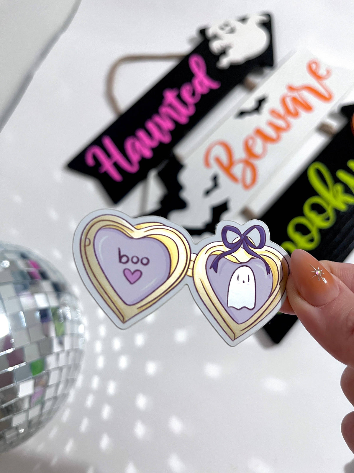 A hand holds a pair of novelty heart-shaped sunglasses with a purple and gold frame, featuring 'boo' on one lens and a ghost on the other. In the blurred background, other Halloween-themed sunglasses are visible, along with a disco ball.