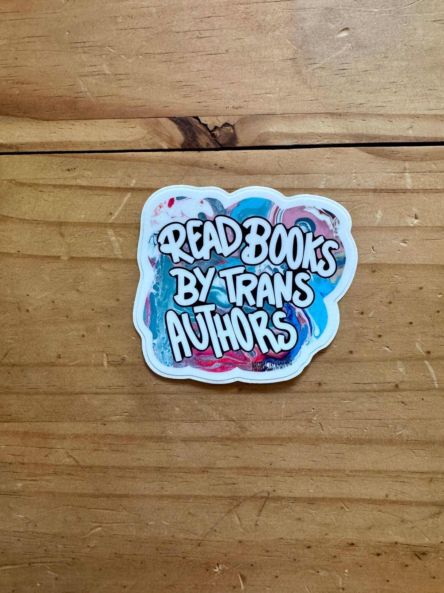 Sticker with 'Read books by trans authors' text on a wooden surface