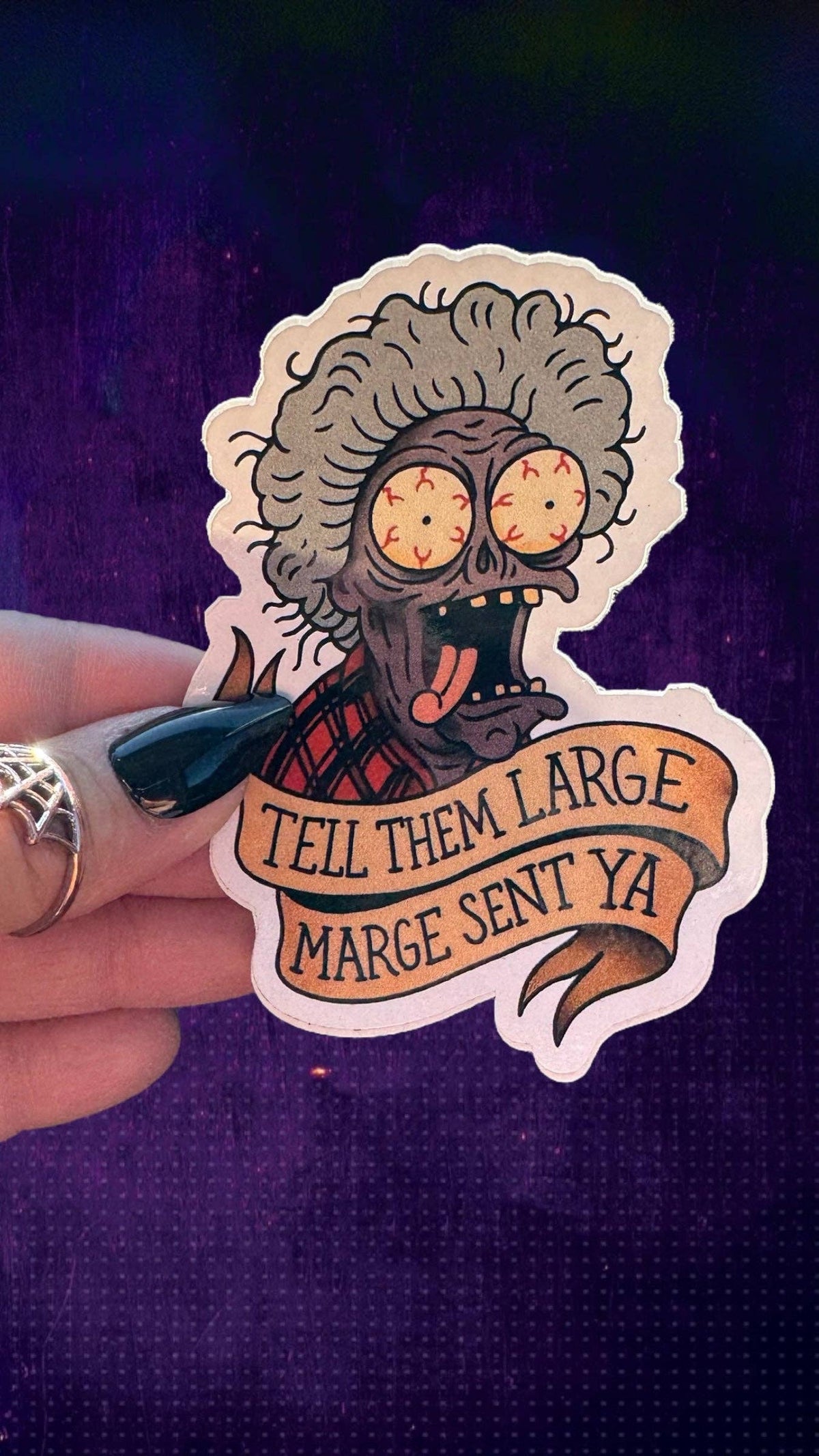 A hand holds a die-cut sticker featuring a cartoon zombie with gray hair, bulging yellow eyes, and a red plaid shirt. The sticker includes a banner reading 'TELL THEM LARGE MARGE SENT YA'.