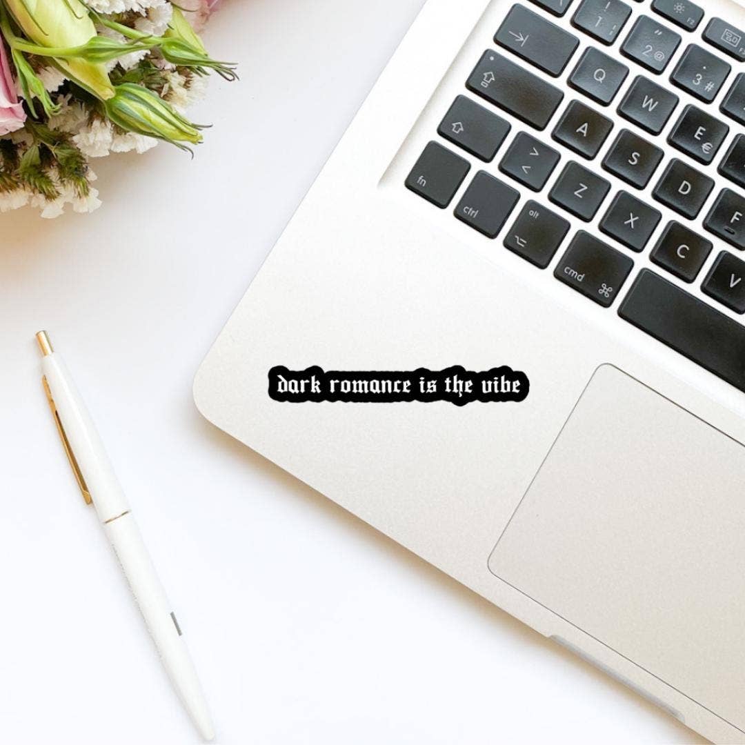 Laptop with 'dark romance is the vibe' sticker on a white surface with flowers.