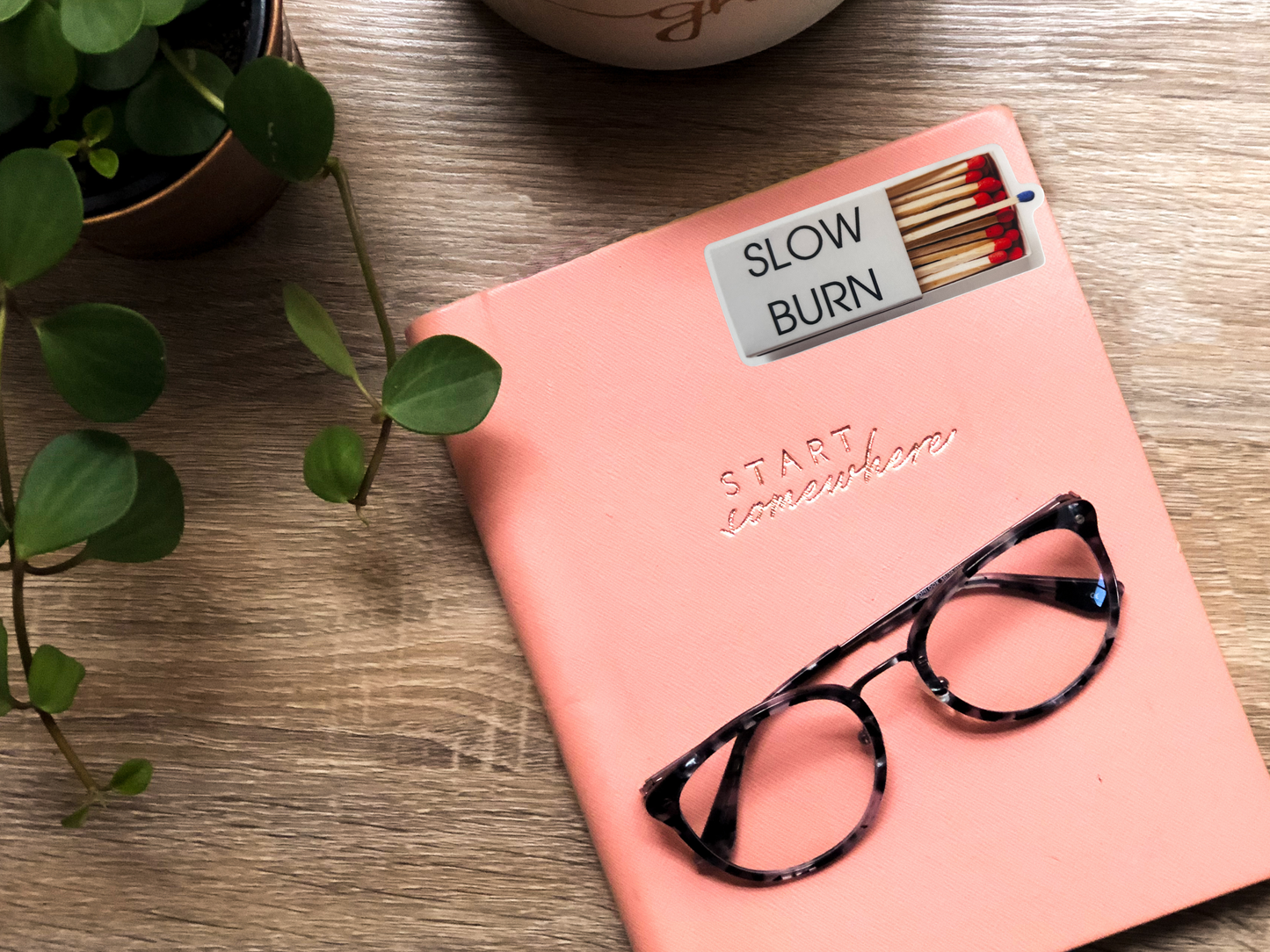 Pink notebook with matches labeled 'Slow Burn' and glasses on a wooden surface.