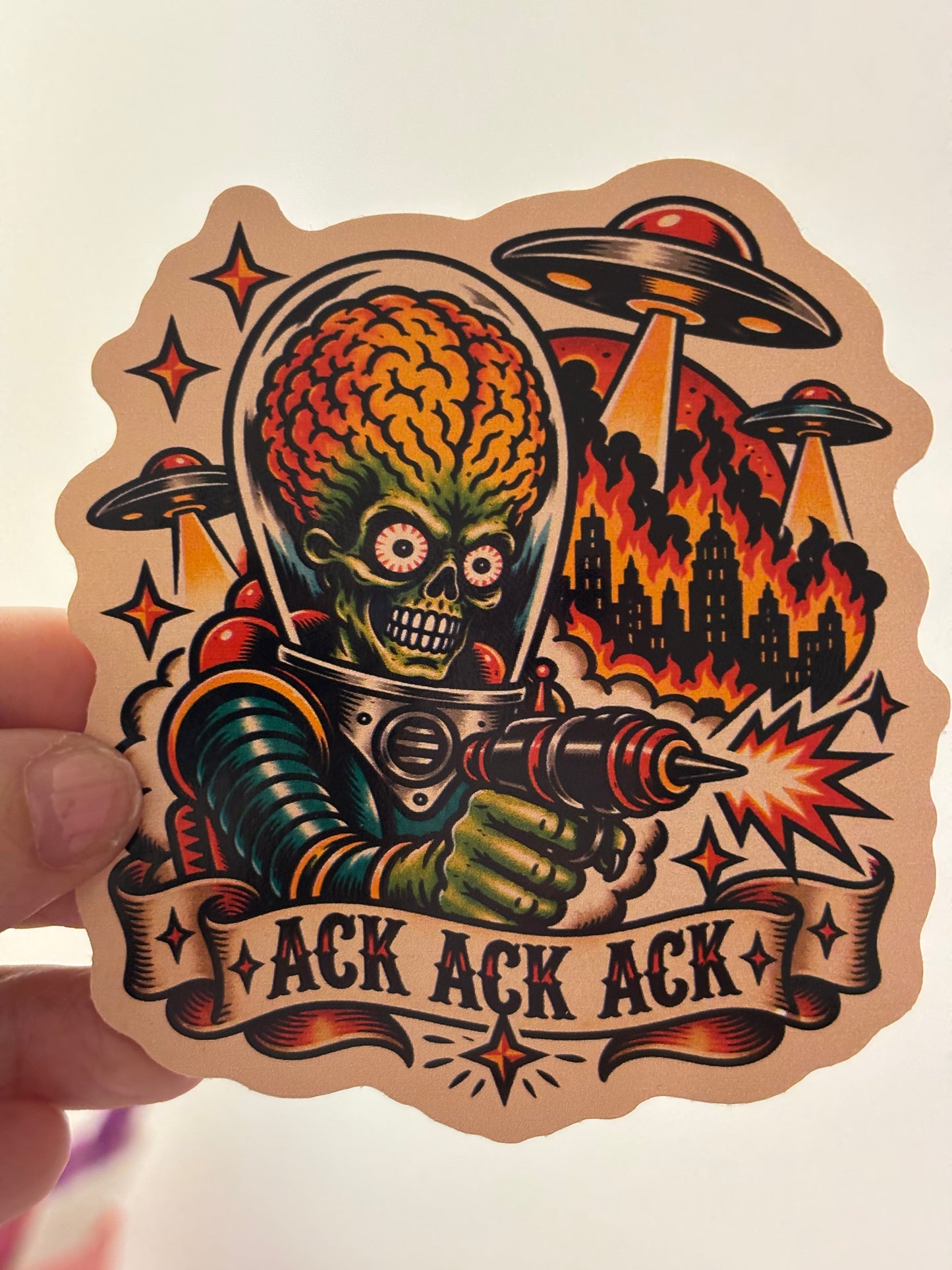 A hand holds a die-cut sticker featuring a retro-style alien-themed illustration with a green skull-headed figure in space gear firing a cannon, surrounded by UFOs, stars, and the text 'ACK ACK ACK' on a banner. The sticker is shown against a plain light background.