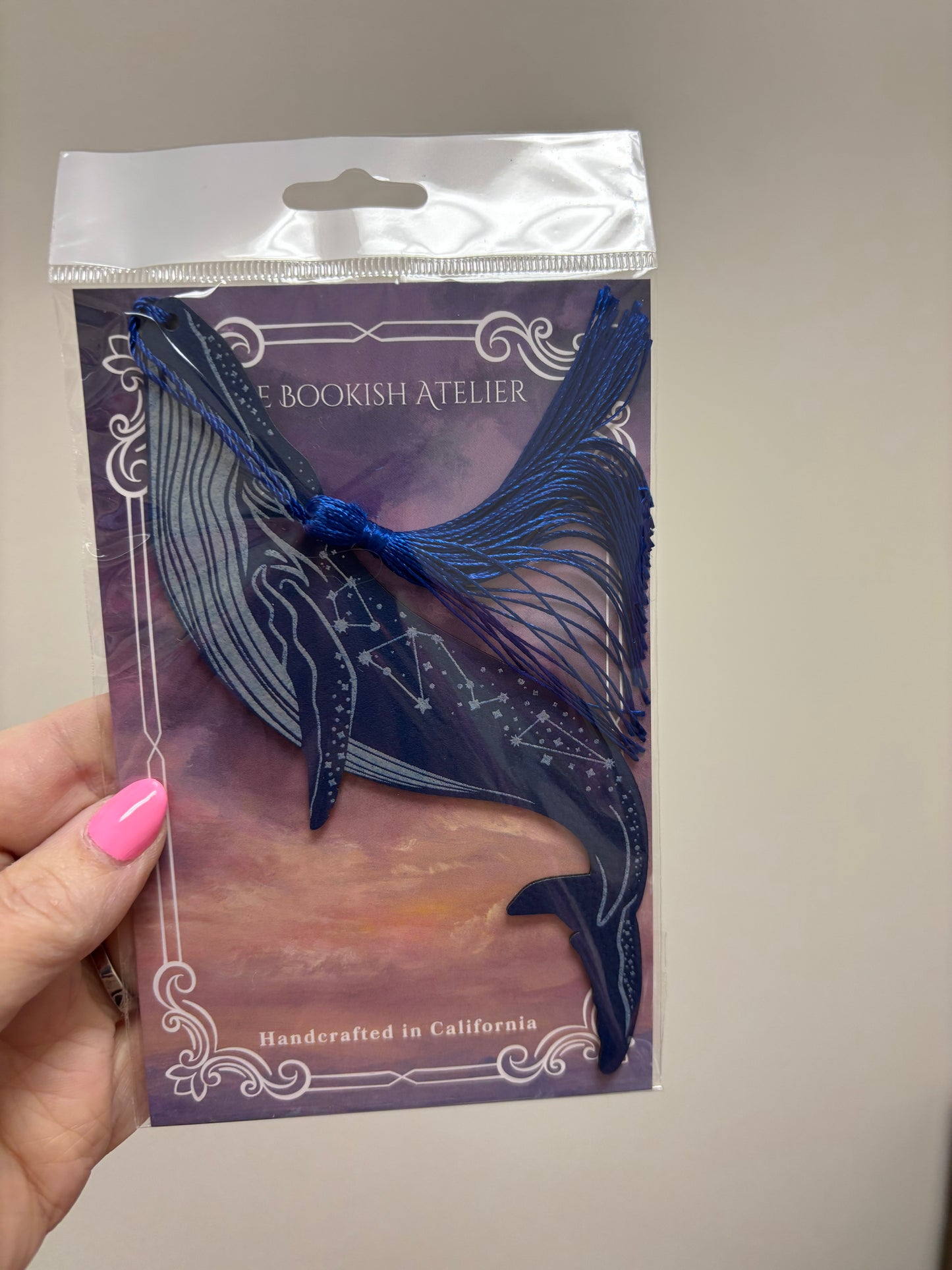 Blue tassled bookmark in clear packaging held by a hand with pink nail polish.