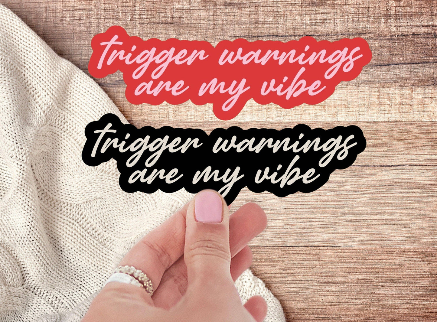 Hand holding two 'trigger warnings are my vibe' stickers on a wooden surface.