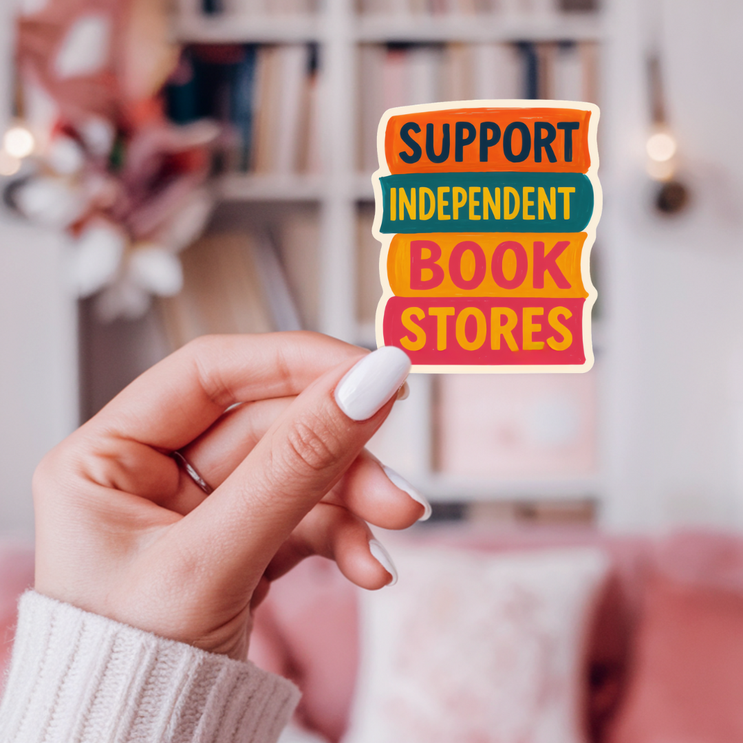 Hand holding a colorful sticker that says 'Support Independent Book Stores' in a blurred indoor setting.