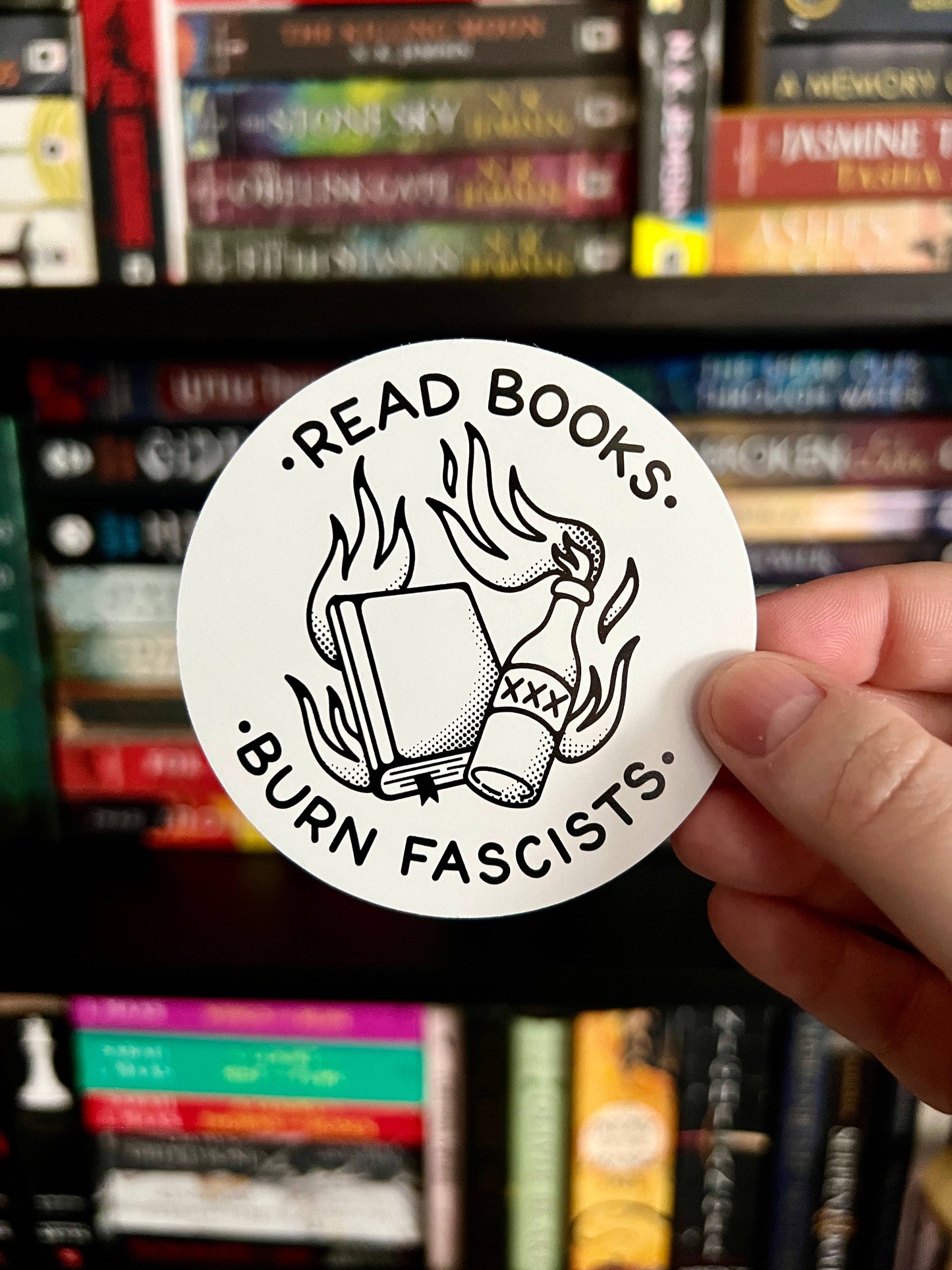 Sticker with 'Read Books, Burn Fascists' design held in front of a bookshelf.