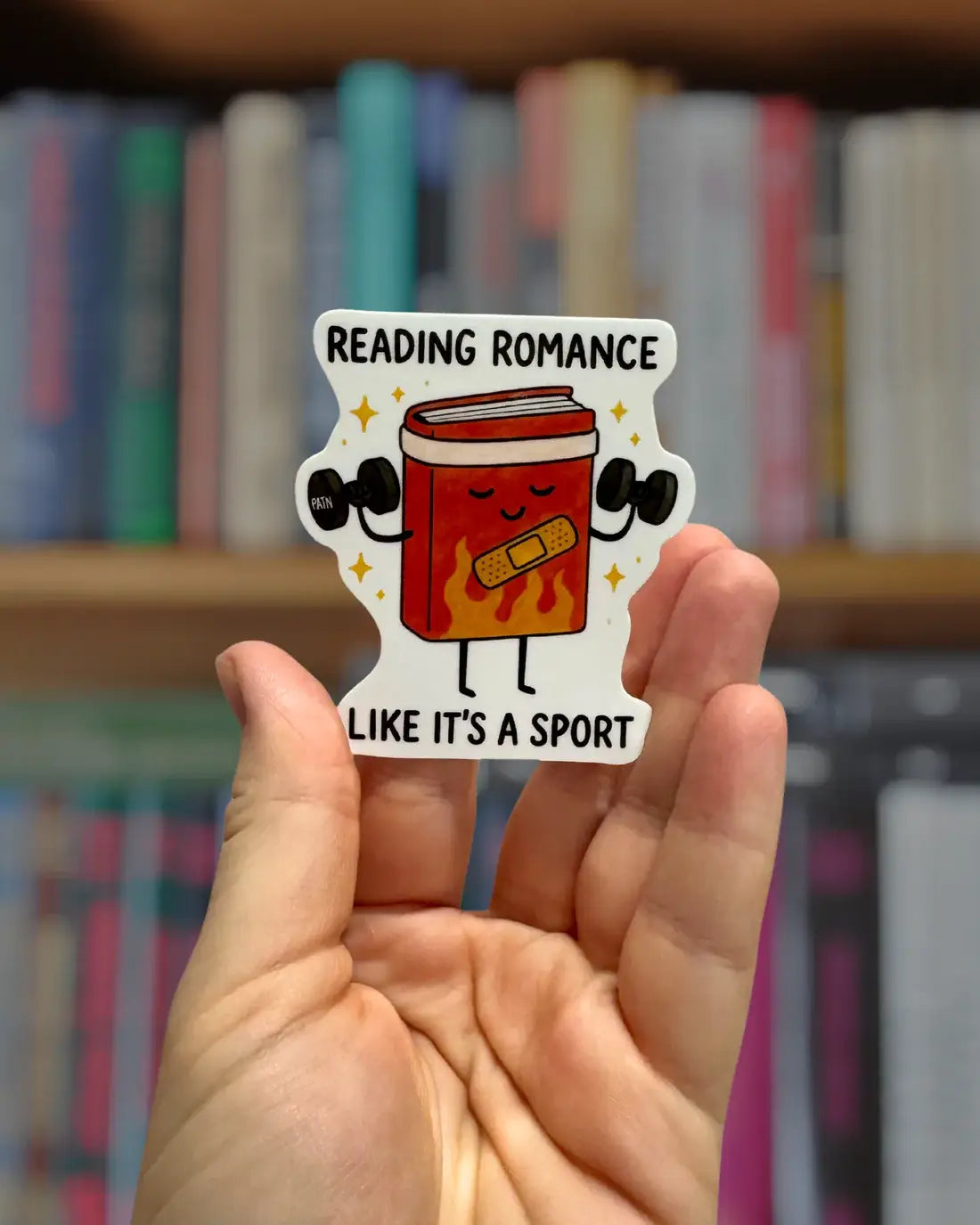 Sticker with a cartoon book character and text 'Reading romance like it's a sport' held in front of a bookshelf.