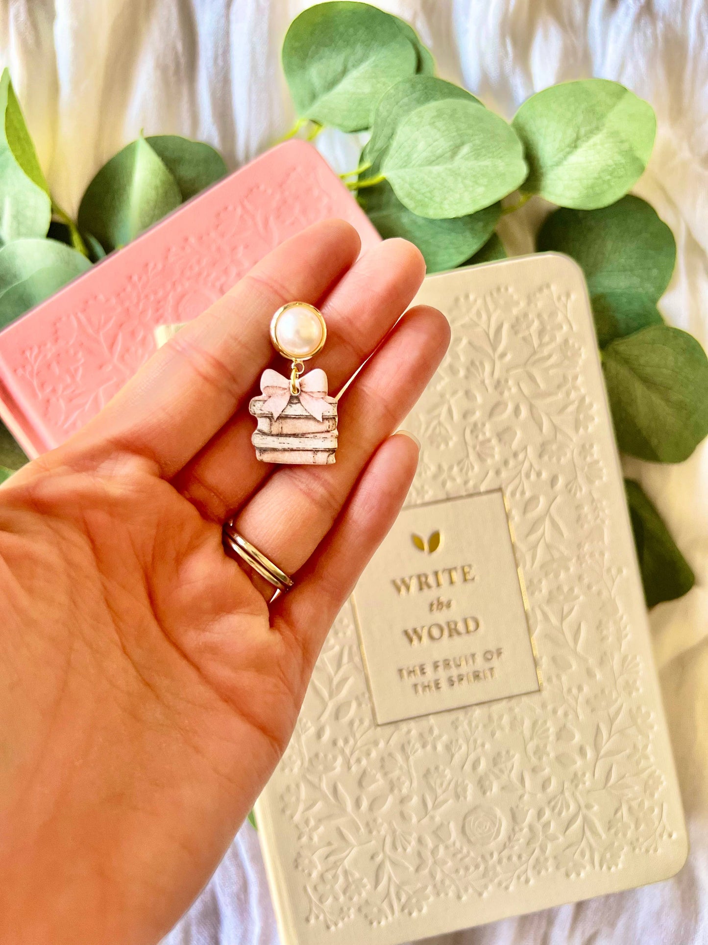 Hand holding a decorative earring with a book and green leaves in the background