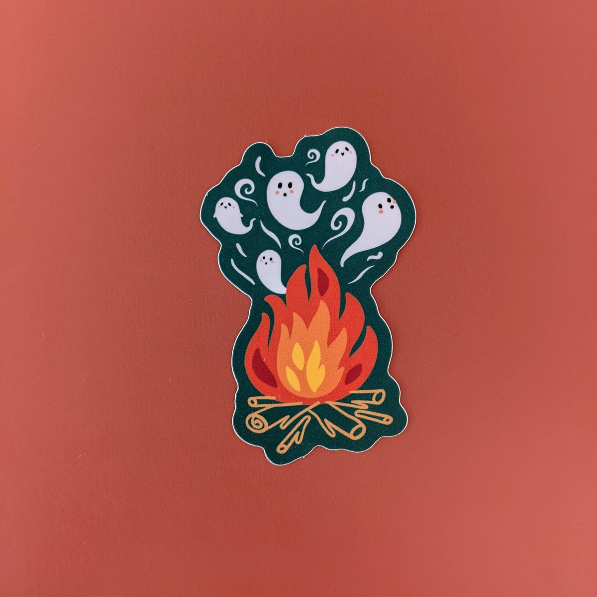 A die-cut sticker featuring a cute cartoon campfire with orange and yellow flames, white ghosts, and a dark teal smoke, shown on a solid terracotta background.