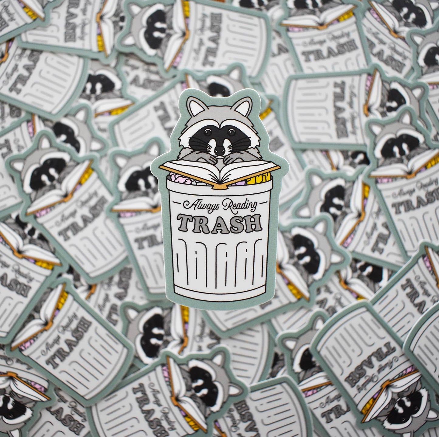 Sticker with a raccoon reading 'Always Reading Trash' on a background of similar stickers.