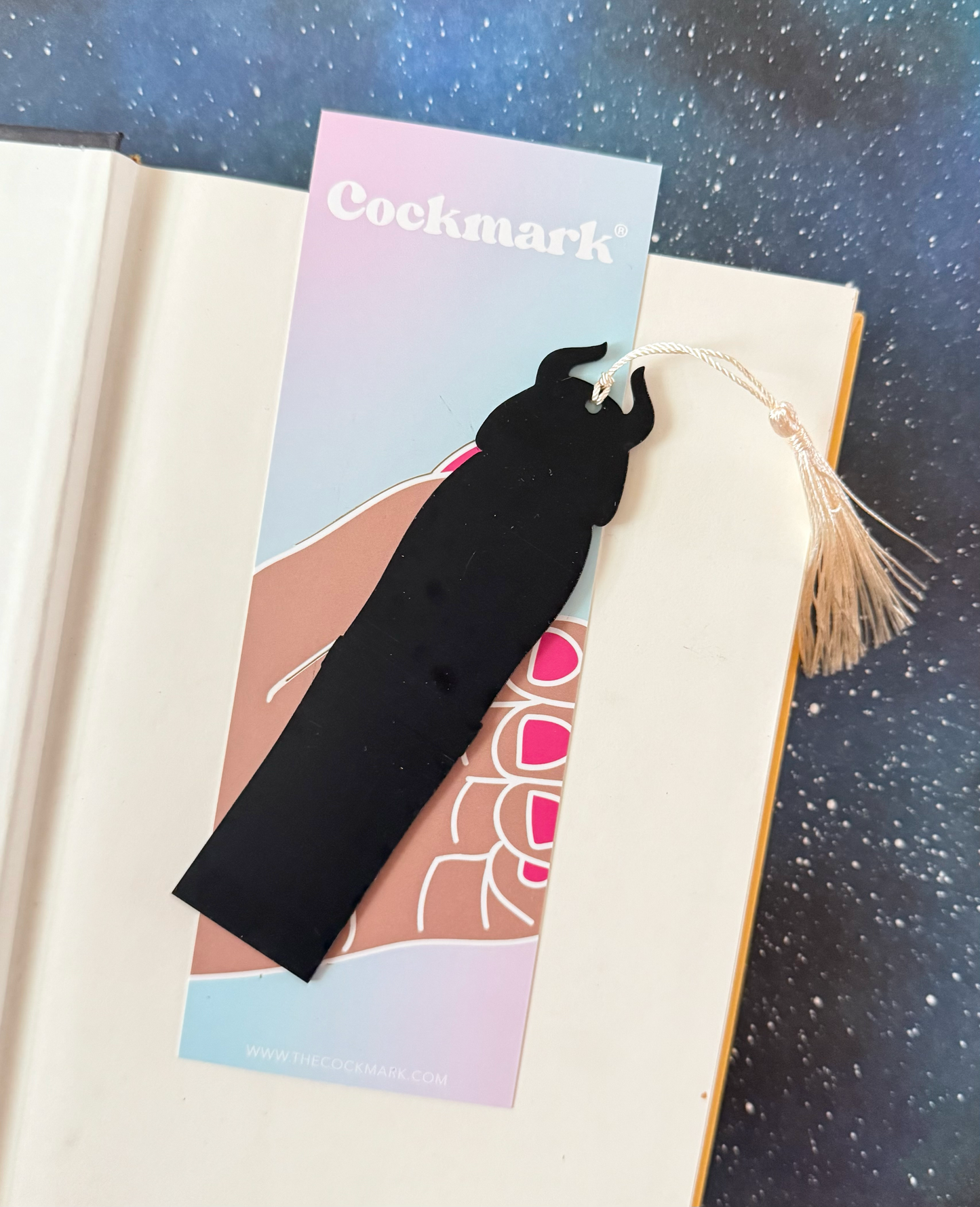 Black tassel bookmark on a colorful card with 'Cockmark' branding, placed on an open book.