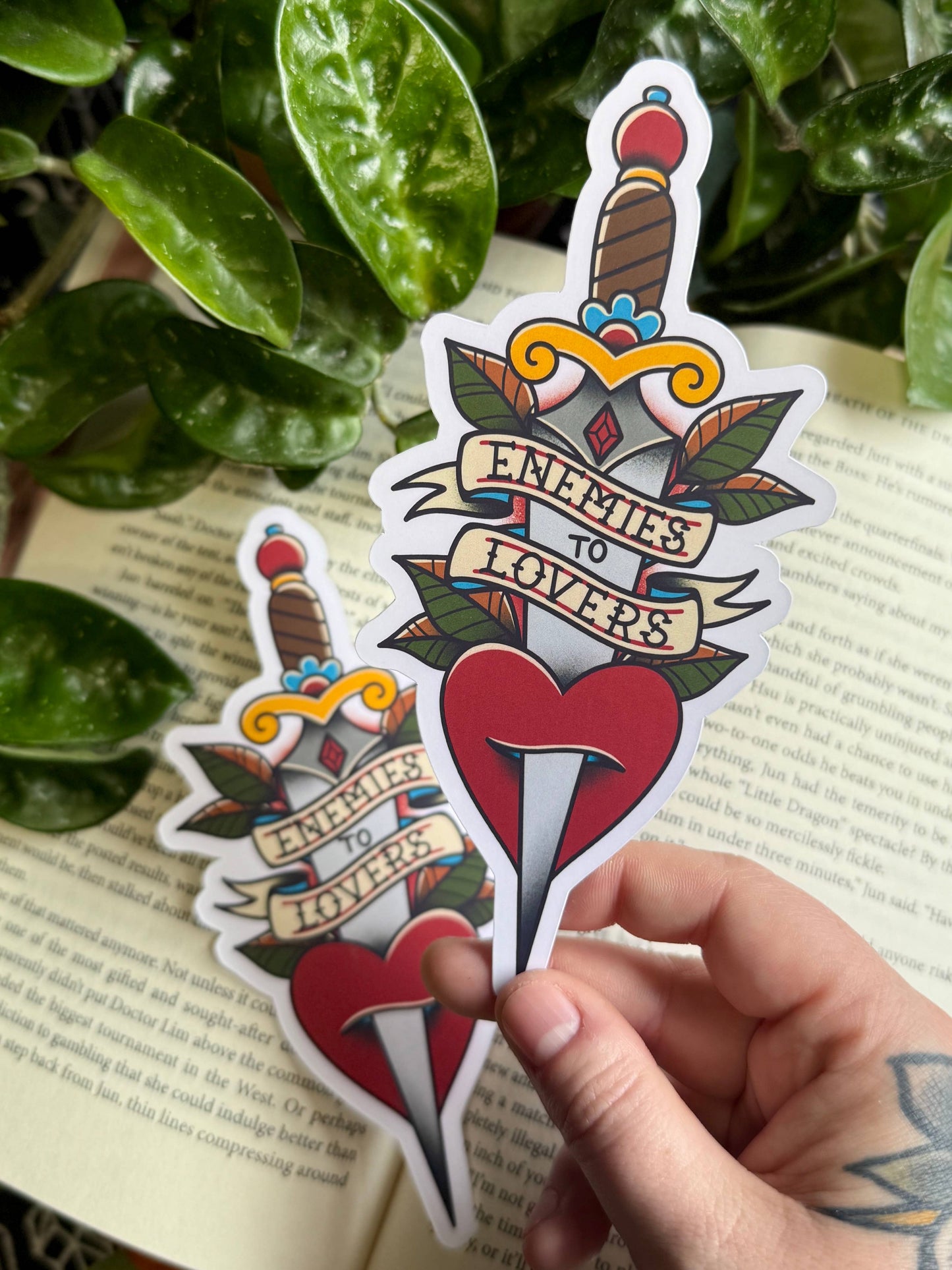 Sticker with a dagger and heart design held by a hand on a book page with leaves in the background