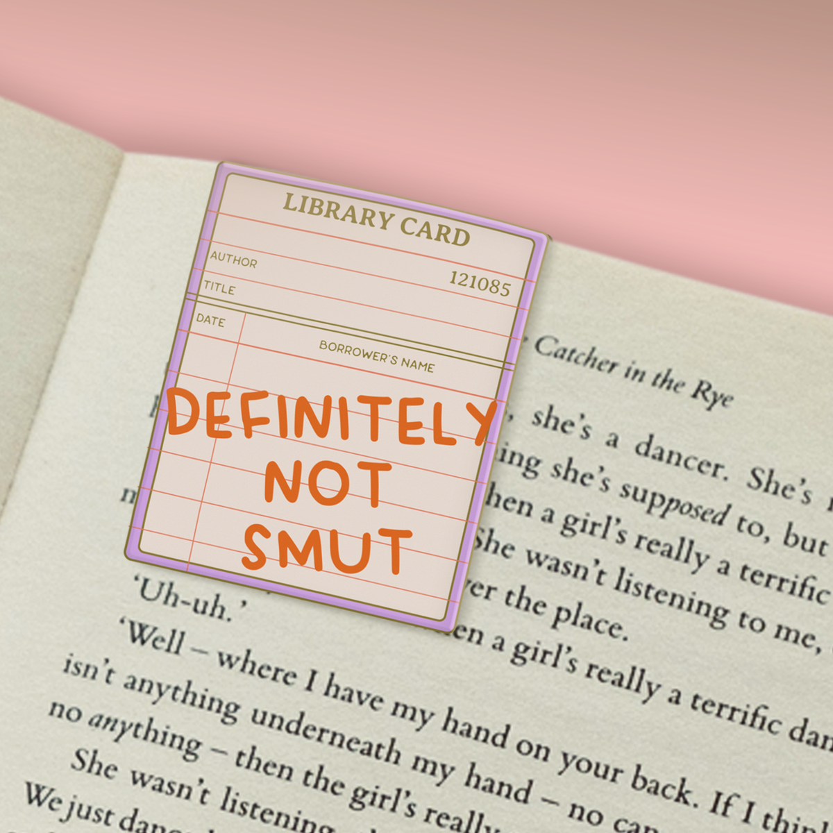 A library card bookmark with the phrase 'DEFINITELY NOT SMUT' written in orange text, placed over an open book page against a pink background.