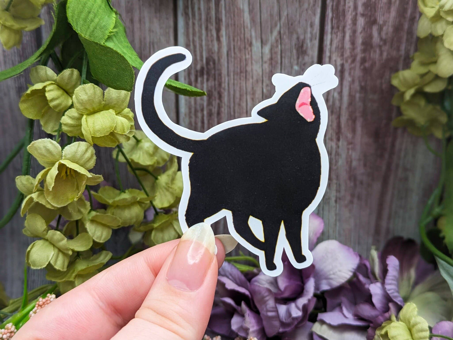 Hand holding a black cat-shaped sticker with flowers and a wooden surface in the background