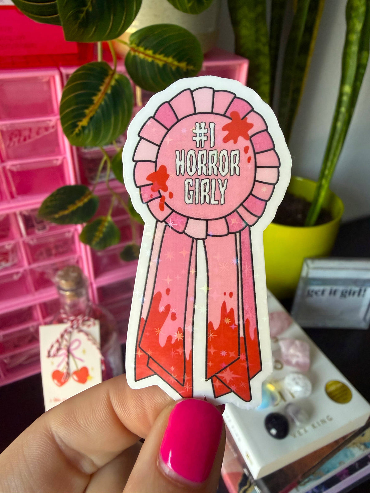A hand with bright pink fingernails holds a die-cut sticker shaped like a pink ribbon award. The sticker reads “#1 HORROR GIRL” in white text, with pink and red horror-themed graphics.