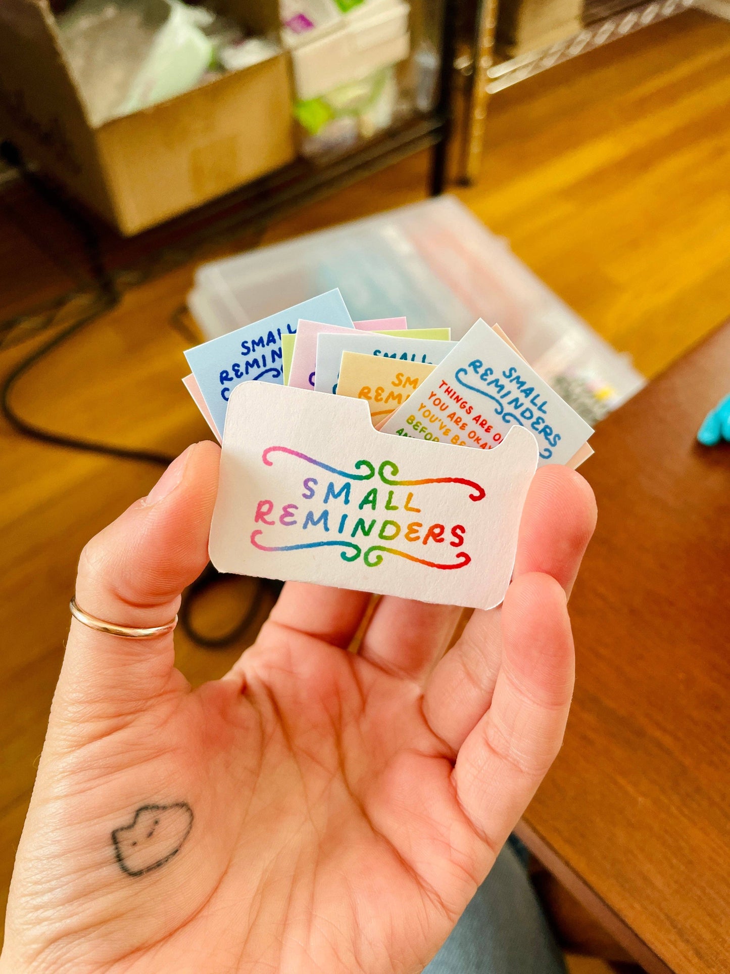 Hand holding a small white pouch with 'Small Reminders' text, surrounded by more colorful pouches on a wooden surface.