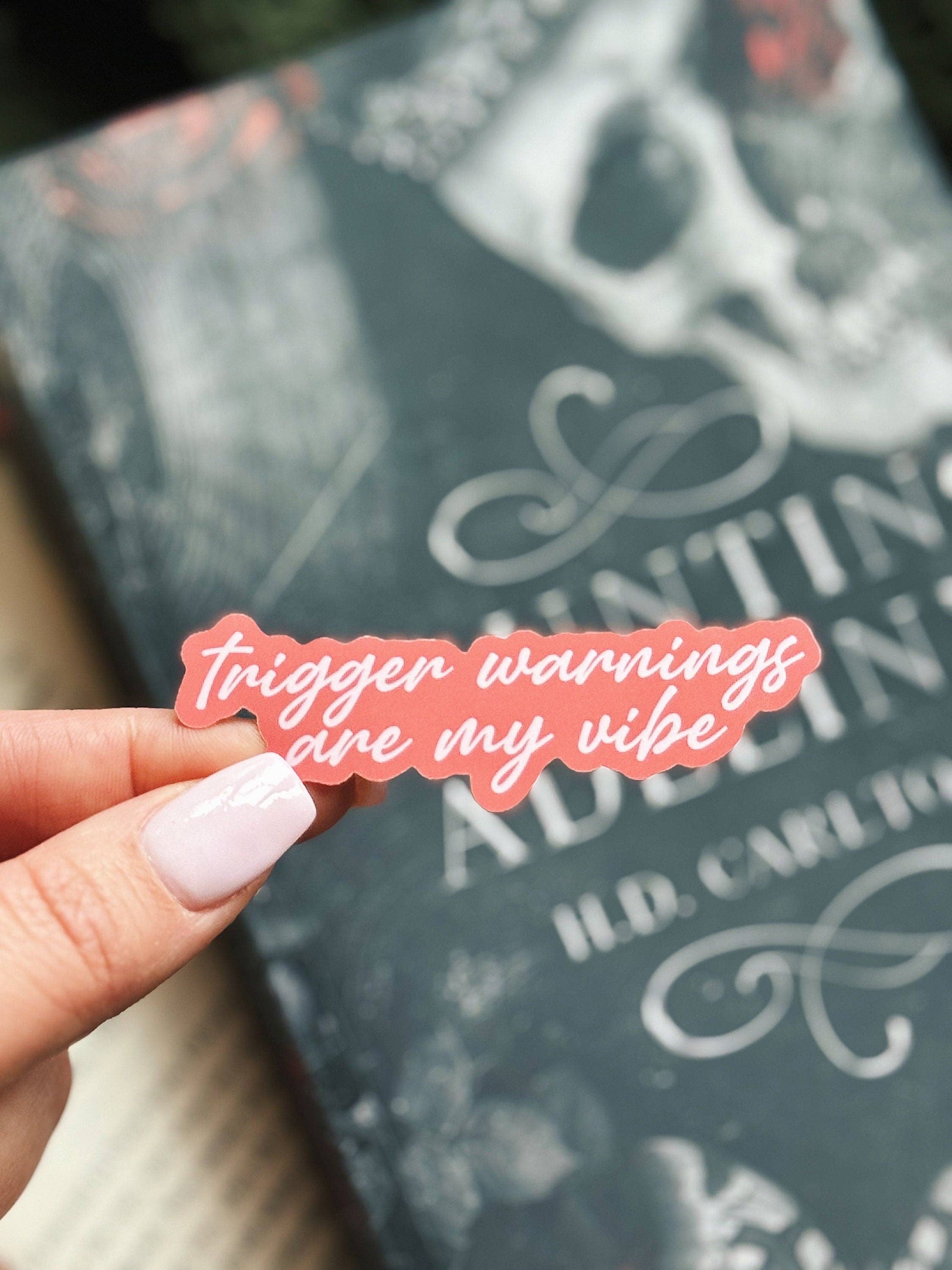 Pink sticker with 'trigger warnings are my vibe' text held over a book