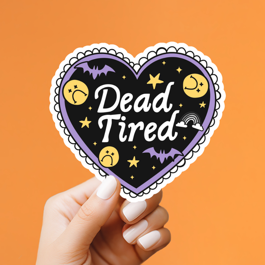 A hand with white manicured nails holds a heart-shaped sticker against a solid orange background. The sticker features a black background with the words 'Dead Tired' in white script, surrounded by purple bats, yellow stars, sad faces, and a small rainbow, all framed by a purple border and a white scalloped edge.