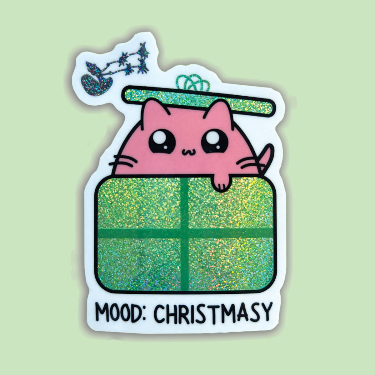 A die-cut sticker featuring a cute pink cartoon cat peeking over a glittery green Christmas gift box, with a small sleigh and stars above and the text 'MOOD: CHRISTSMASY' at the bottom, shown on a pale green background.