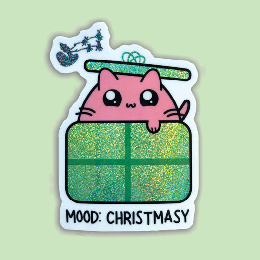 A die-cut sticker featuring a cute pink cartoon cat peeking over a glittery green Christmas gift box, with a small sleigh and stars above and the text 'MOOD: CHRISTSMASY' at the bottom, shown on a pale green background.