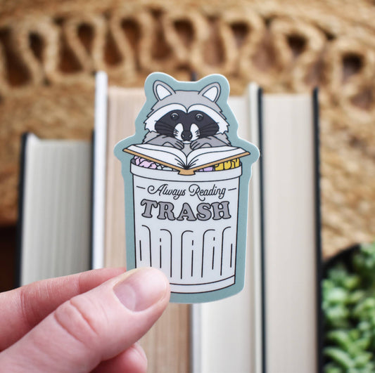 Sticker of a raccoon in a trash can with 'Always Reading Trash' text, held by a hand.