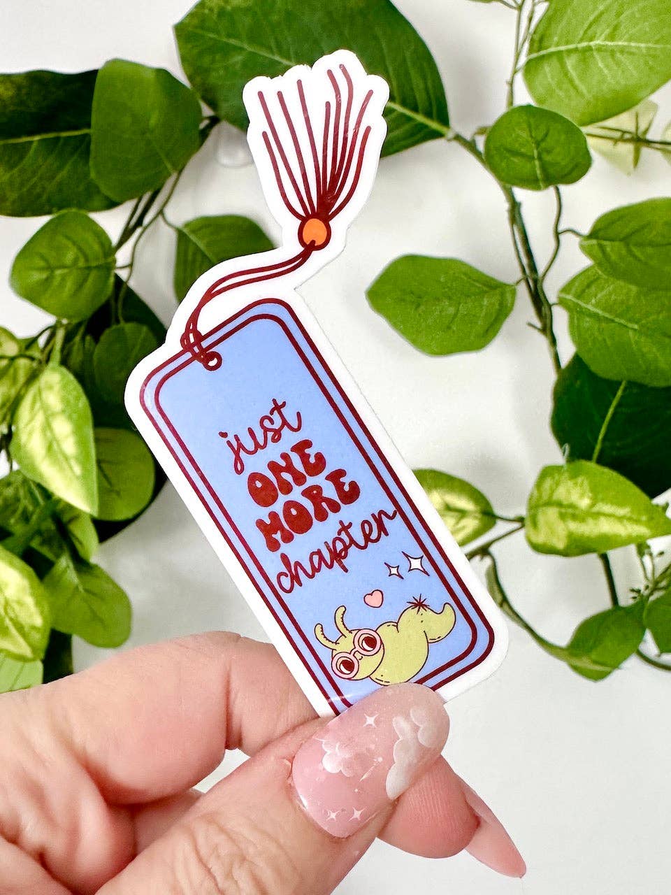 Sticker with 'Just One More Chapter' text held by a hand against a green leafy background