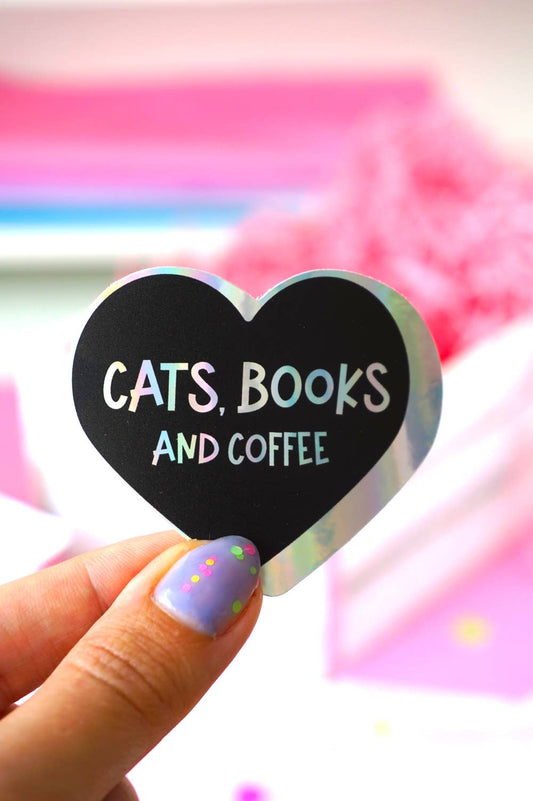 A hand with lavender and green glitter nail polish holds a heart-shaped black sticker that reads 'CATS, BOOKS AND COFFEE' in white, pink, and blue lettering. The background is a soft-focus pink and white.