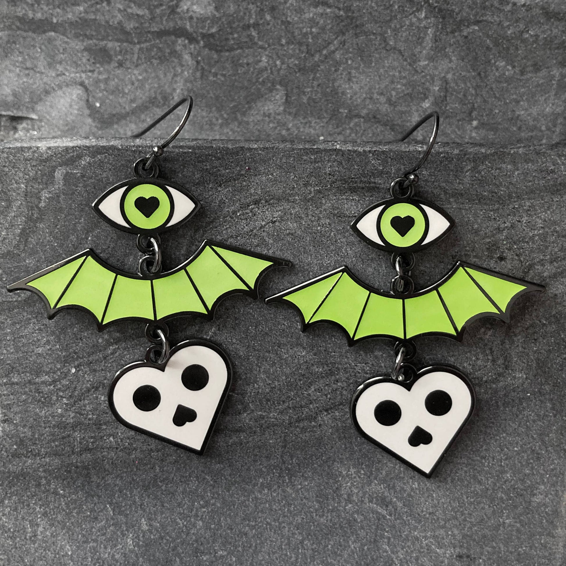 A pair of gothic-style bat dangle earrings with bright green wings, a green eye with a black heart, and a white skull heart charm, displayed on a dark gray textured background.