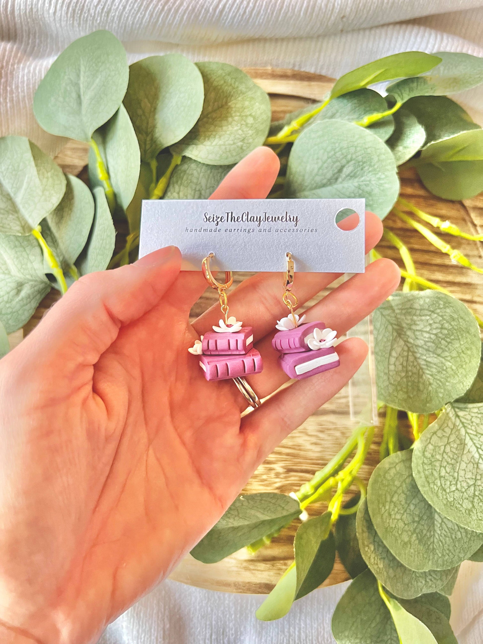 Hand holding a card with earrings featuring stacked books against a leafy background