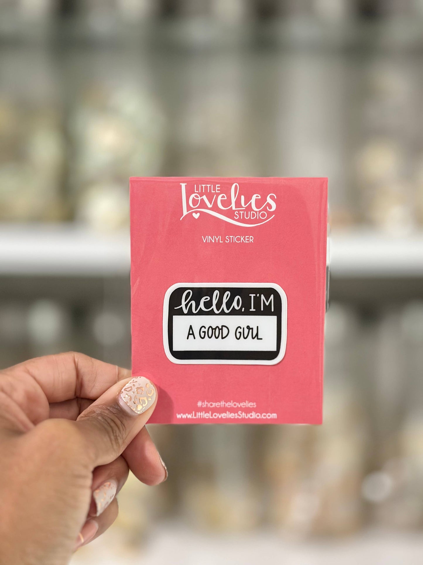 Pink vinyl sticker with 'hello, I'm a good girl' text held by a hand against a blurred background.