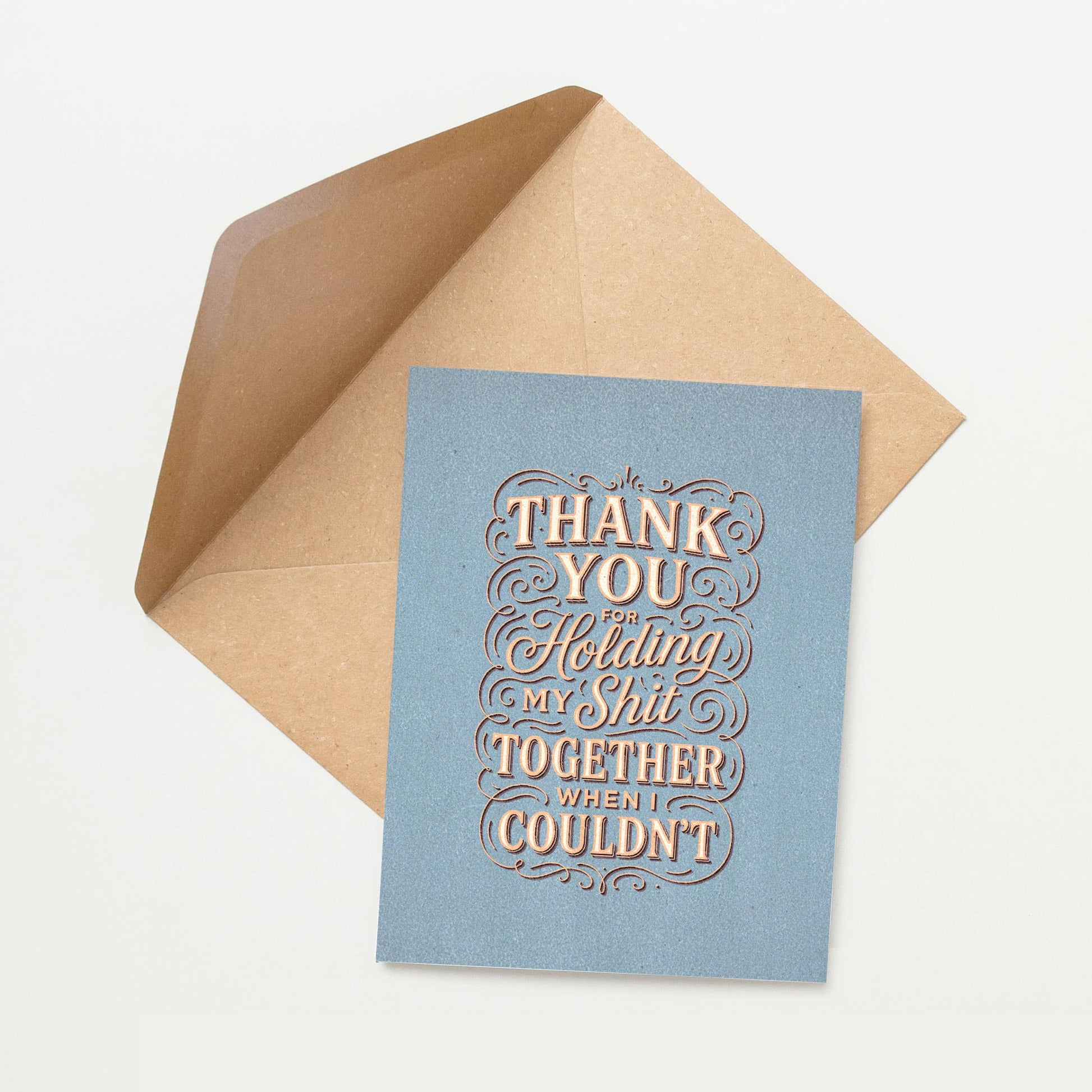 Blue card with gold text 'Thank you for holding my shit together when I couldn't' inside a brown envelope on a white background.