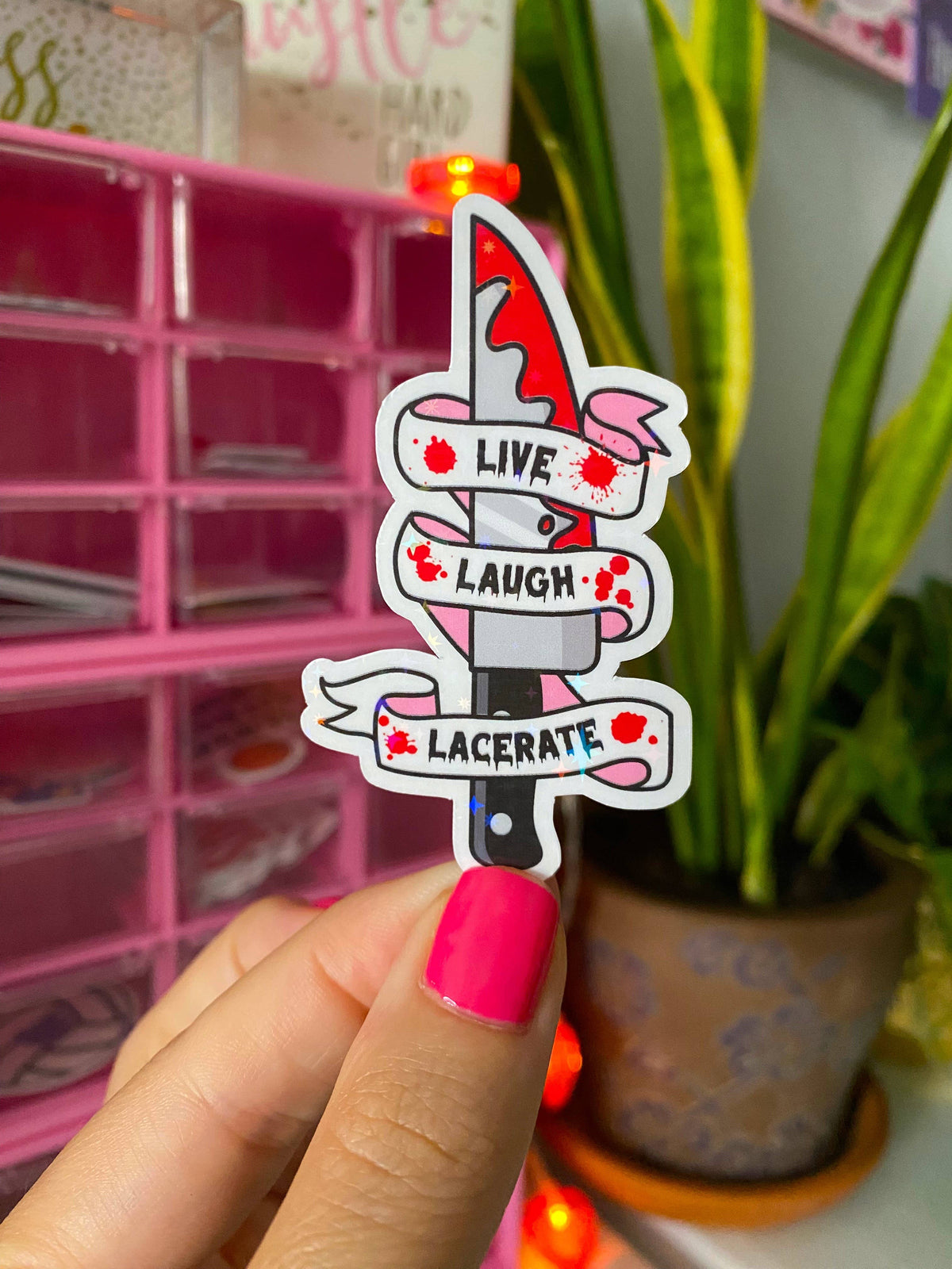 A hand with bright pink nail polish holds a die-cut sticker featuring a red-and-white knife with banners reading “LIVE,” “LAUGH,” and “LACERATE,” against a blurred pink organizer and green plant background.