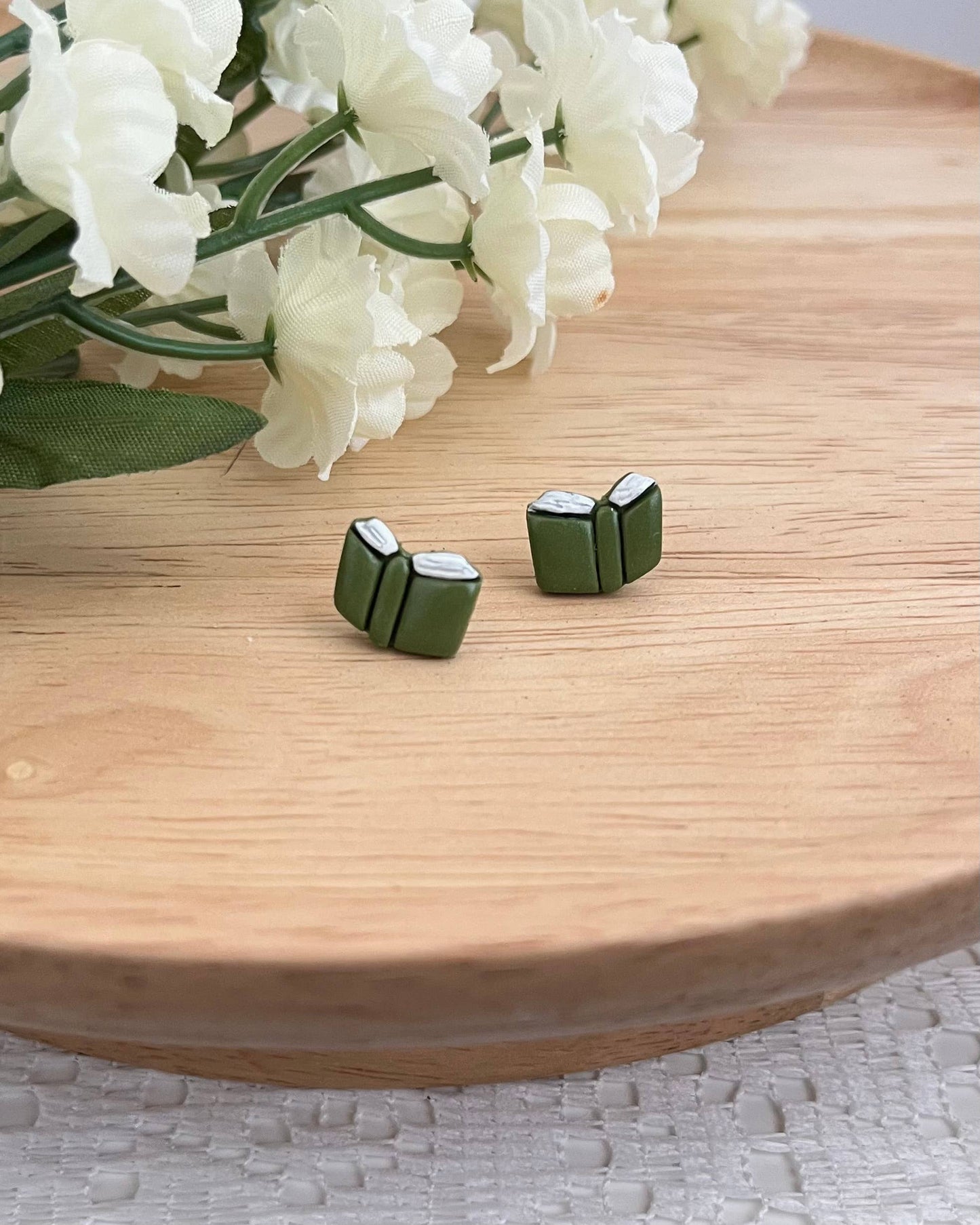 Green book-shaped earrings on a wooden surface with white flowers in the background