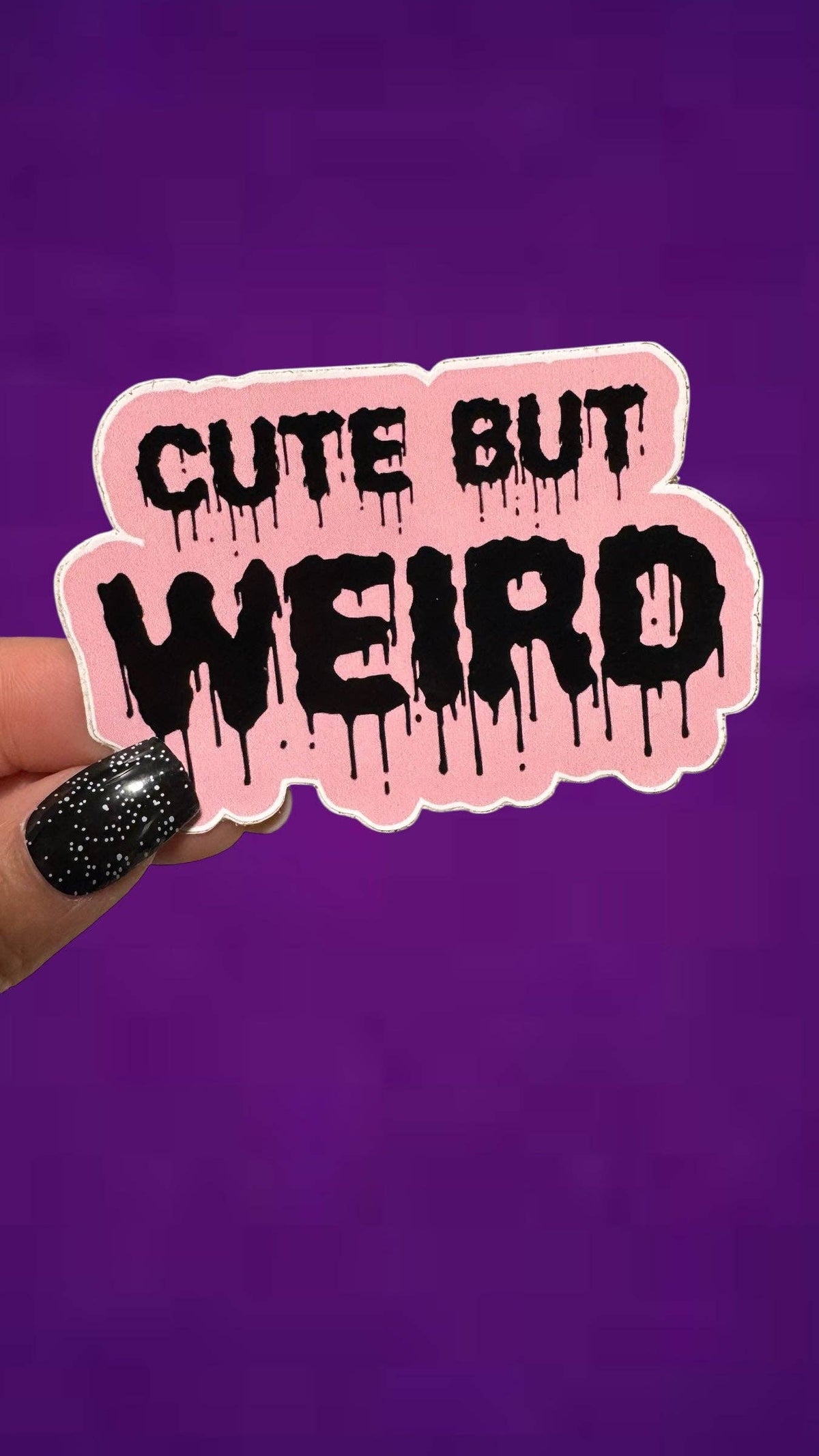 A hand with black glitter nail polish holds a die-cut sticker that reads 'CUTE BUT WEIRD' in dripping black letters on a pink background with a white outline, set against a solid purple backdrop.