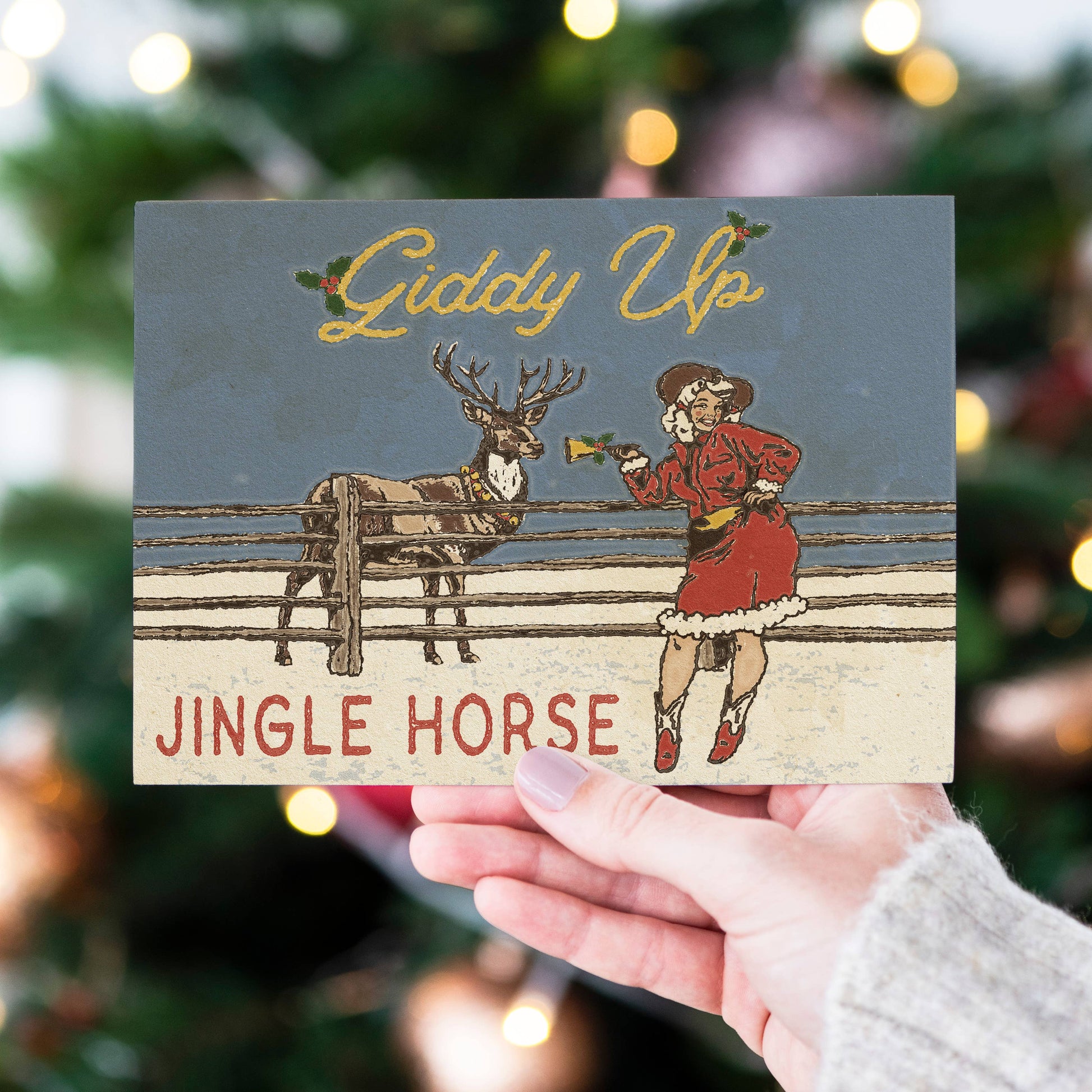 A hand holds a vintage-style Christmas card featuring the text 'Giddy Up' and 'JINGLE HORSE' with an illustration of a person in a red Santa outfit feeding a jingle bell to a reindeer in a snowy scene. The background shows a blurred Christmas tree with warm lights.