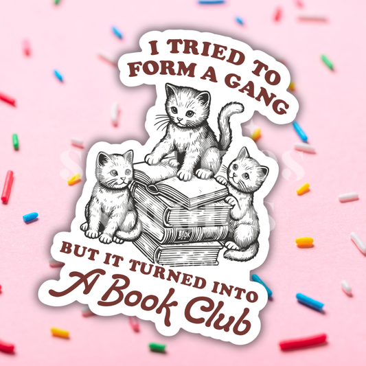 Sticker with cats and text 'I tried to form a gang but it turned into a book club' on a pink background with sprinkles.