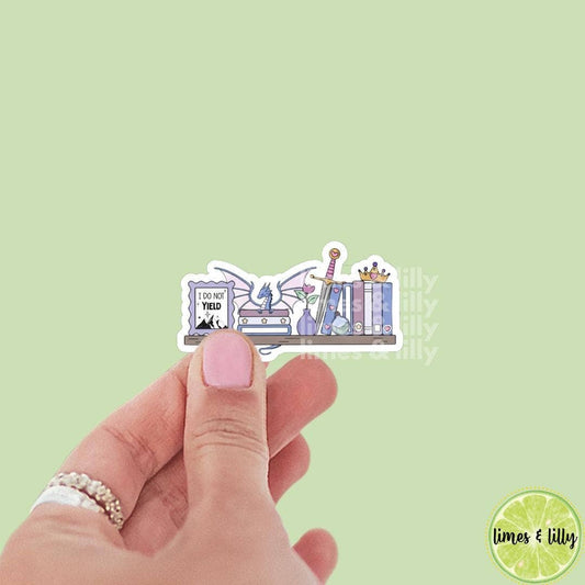 Hand holding a sticker with a design of books and a dragon on a light green background, with 'limes & lilly' branding.