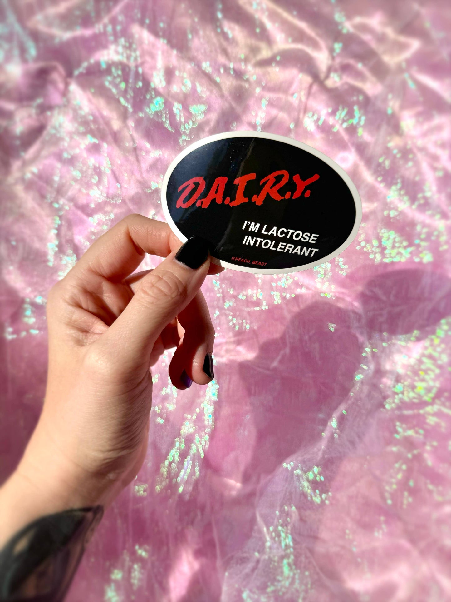 Hand holding a sticker with 'DAIRY I'M LACTOSE INTOLERANT' text against a pink, glittery background.