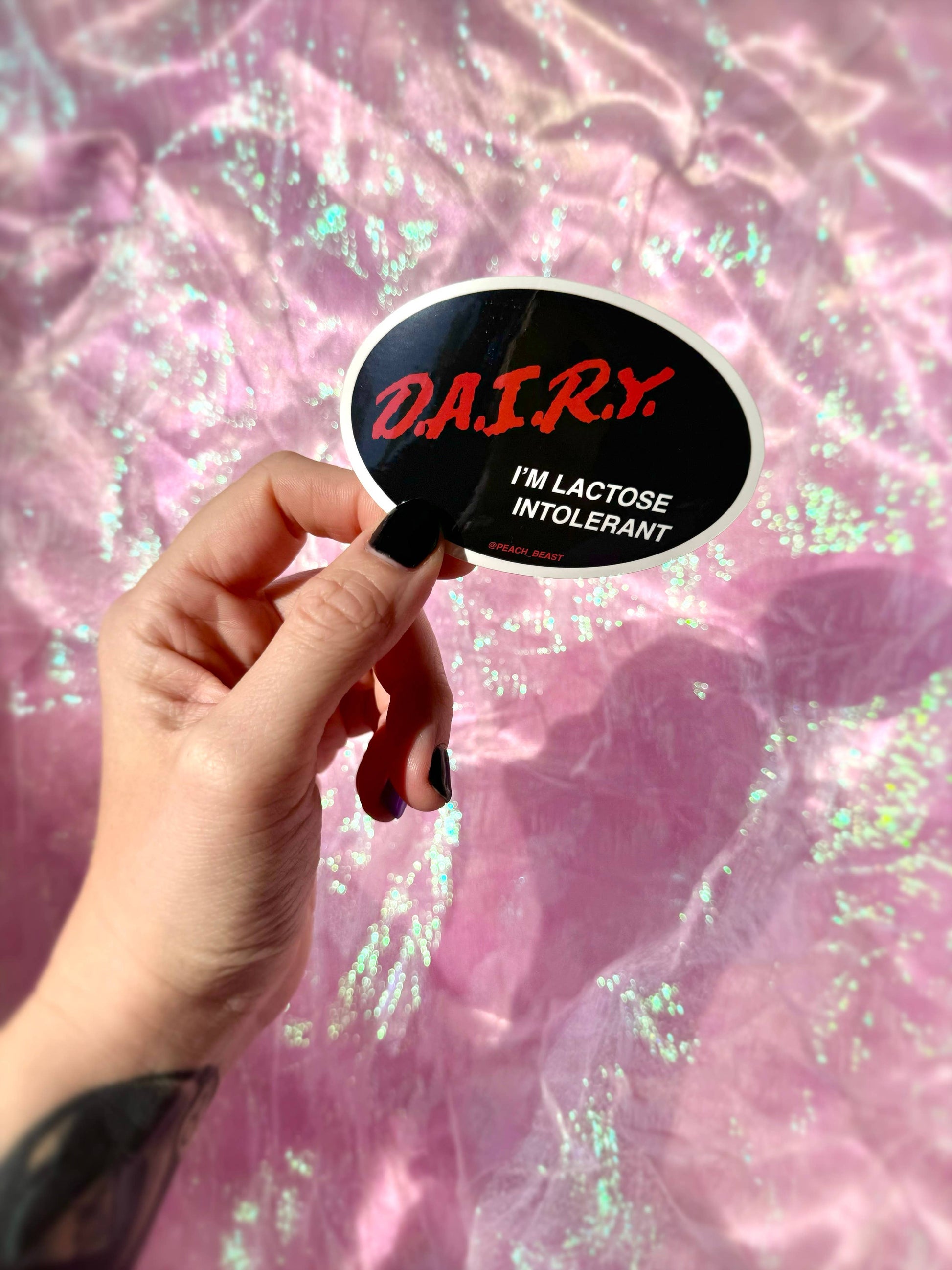 Hand holding a sticker with 'DAIRY I'M LACTOSE INTOLERANT' text against a pink, glittery background.