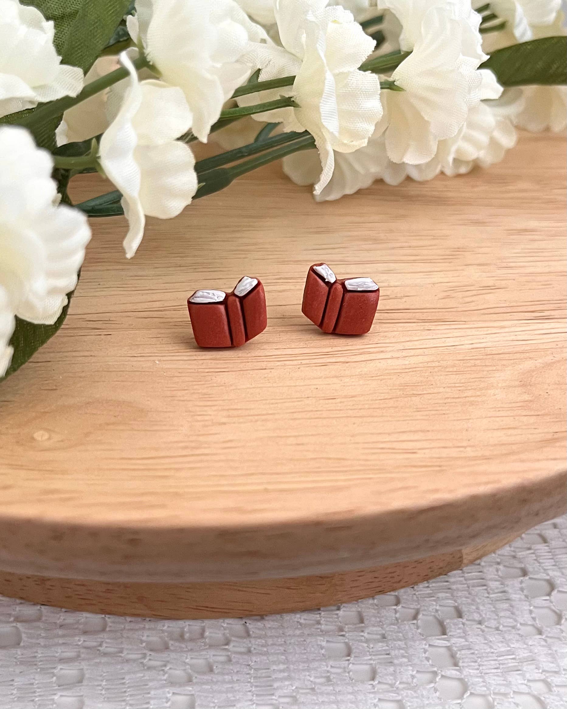 Red book-shaped earrings on a wooden surface with white flowers in the background