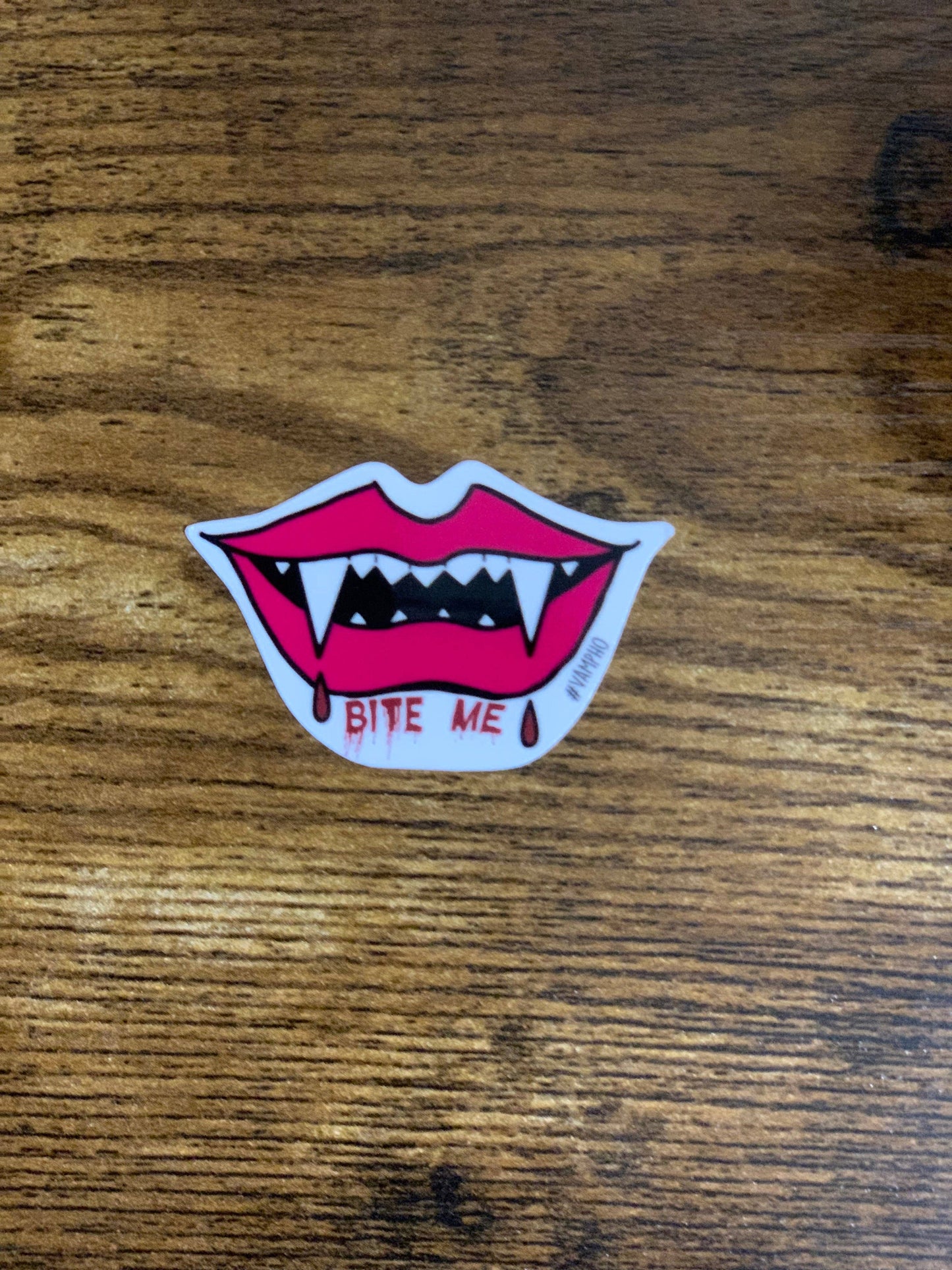 Sticker of a mouth with fangs and 'Bite Me' text on a wooden surface